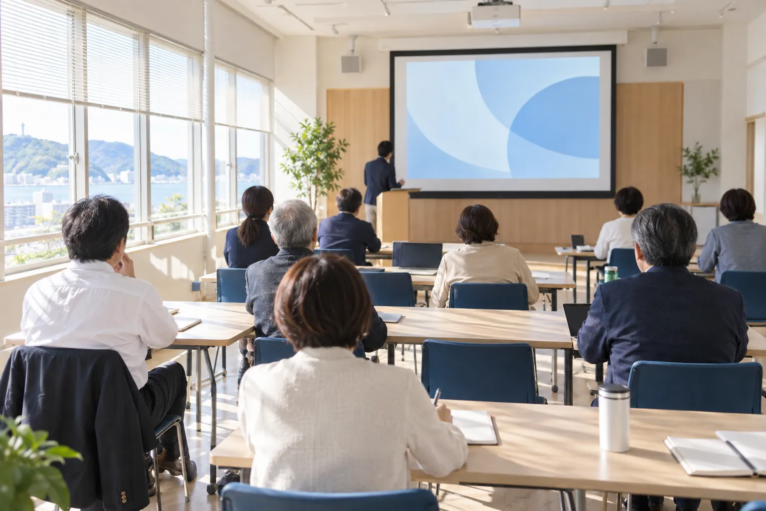 出張IT講座のイメージ。公民館の会議室で、参加者が前のスクリーンを見ながら講師の話を聞いている（後ろ姿）